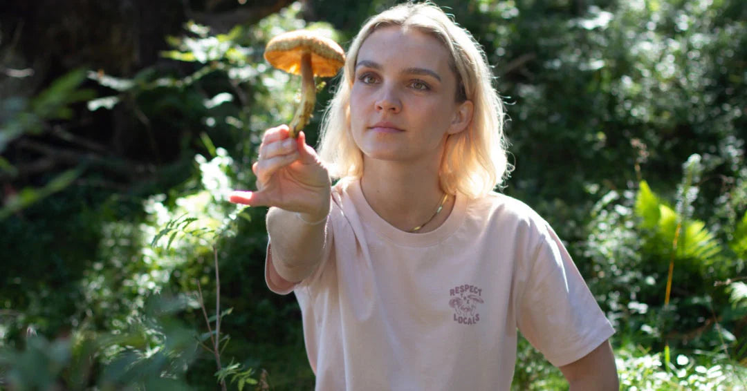 Blonde Frau mit NIKIN Mushroom Shirt schaut auf einen großen Pilz in ausgestreckter Hand.