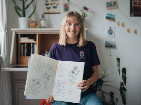 Sophia Hultsch sitzt in ihrem Atelier, lächelt in die Kamera und hält ein geöffnetes Skizzenbuch mit floralen Entwürfen in der Hand. Sie trägt ein violettes NIKIN TreeShirt mit dem „Connected by Nature“-Design.